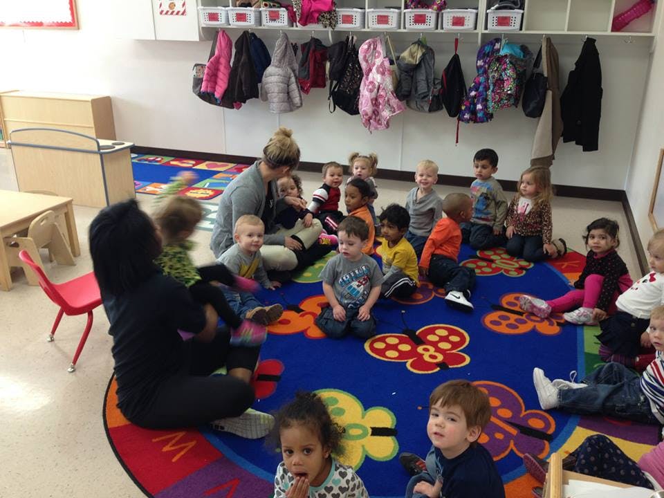 Two people sitting on a rug with a group of children in a classroom