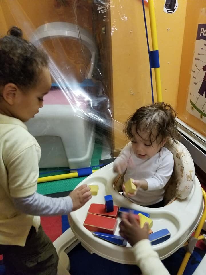 Child in a high chair playing with blocks, another child reaching for a block
