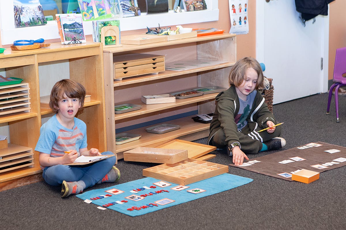 Two children sitting on the floor, one writing and one playing with cards