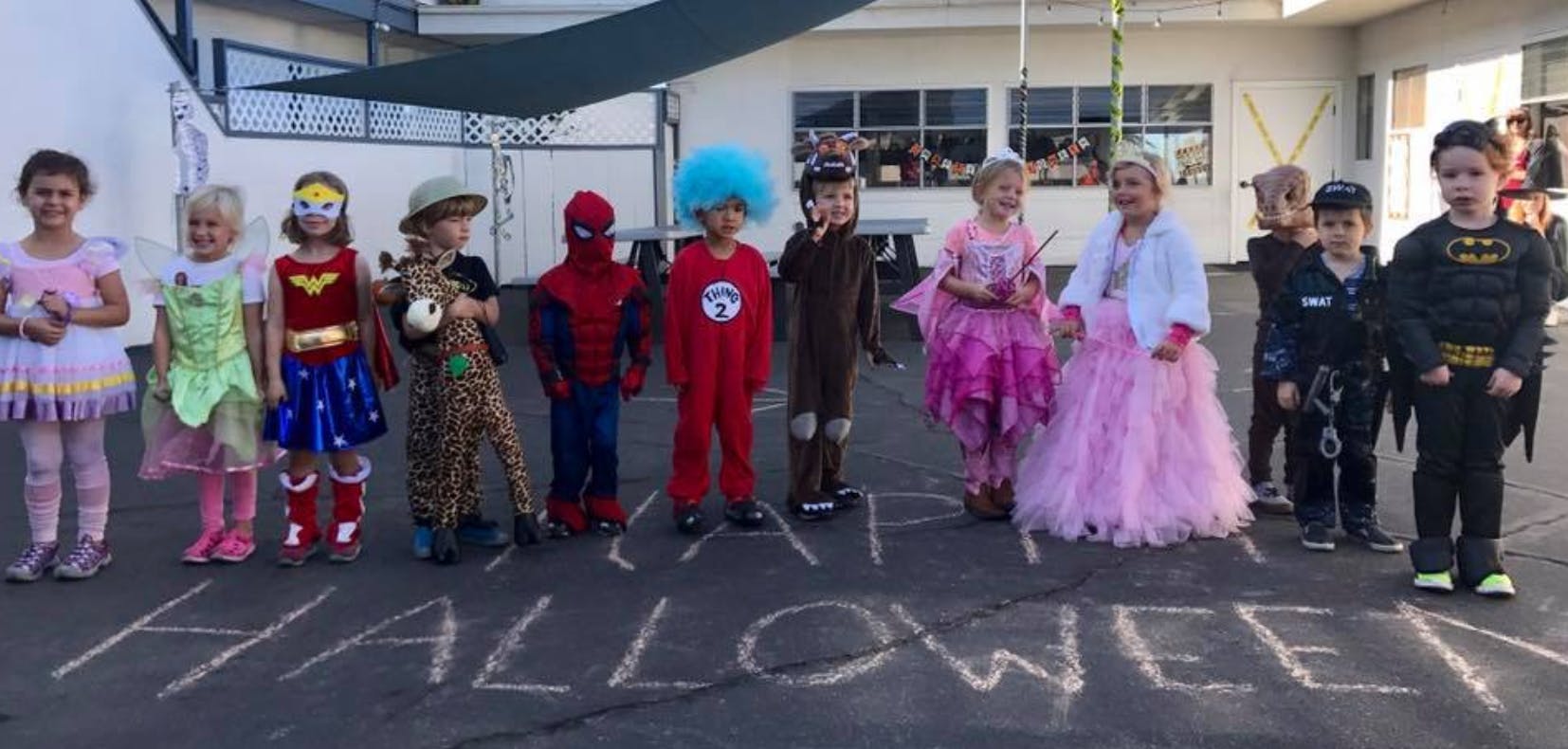 Children in various costumes standing on pavement with "HALLOWEEN" written in chalk
