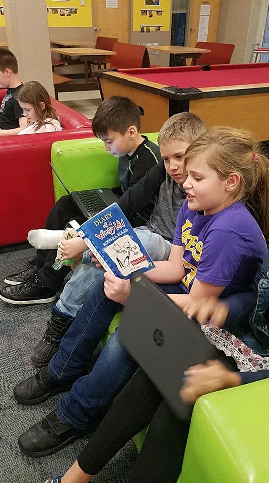 Three children sitting on chairs, one reading a book, another looking at a laptop