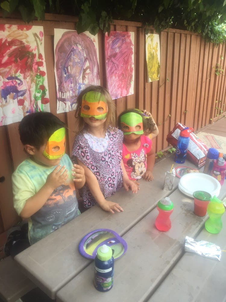 Three children wearing masks standing at a picnic table outdoors