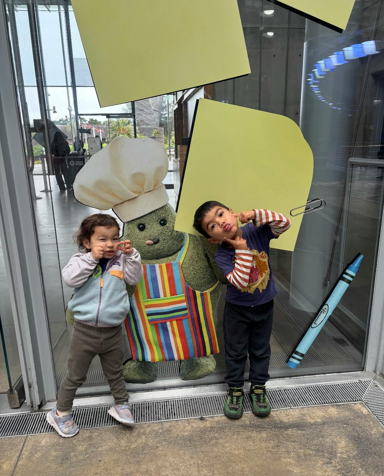 Two children standing and making faces next to a cartoon chef mascot display