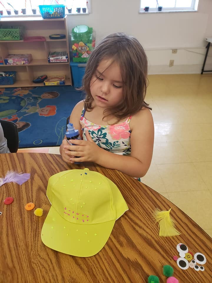 Child holding a bottle of glue over a yellow hat on a table