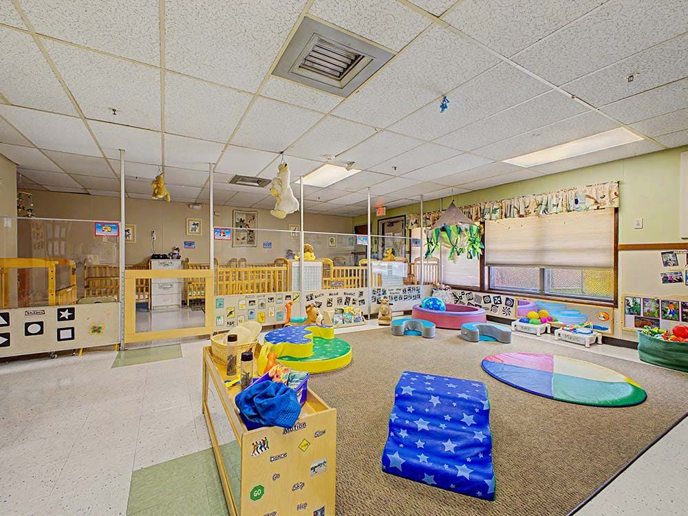 A colorful childcare classroom with cribs, play mats, and toys