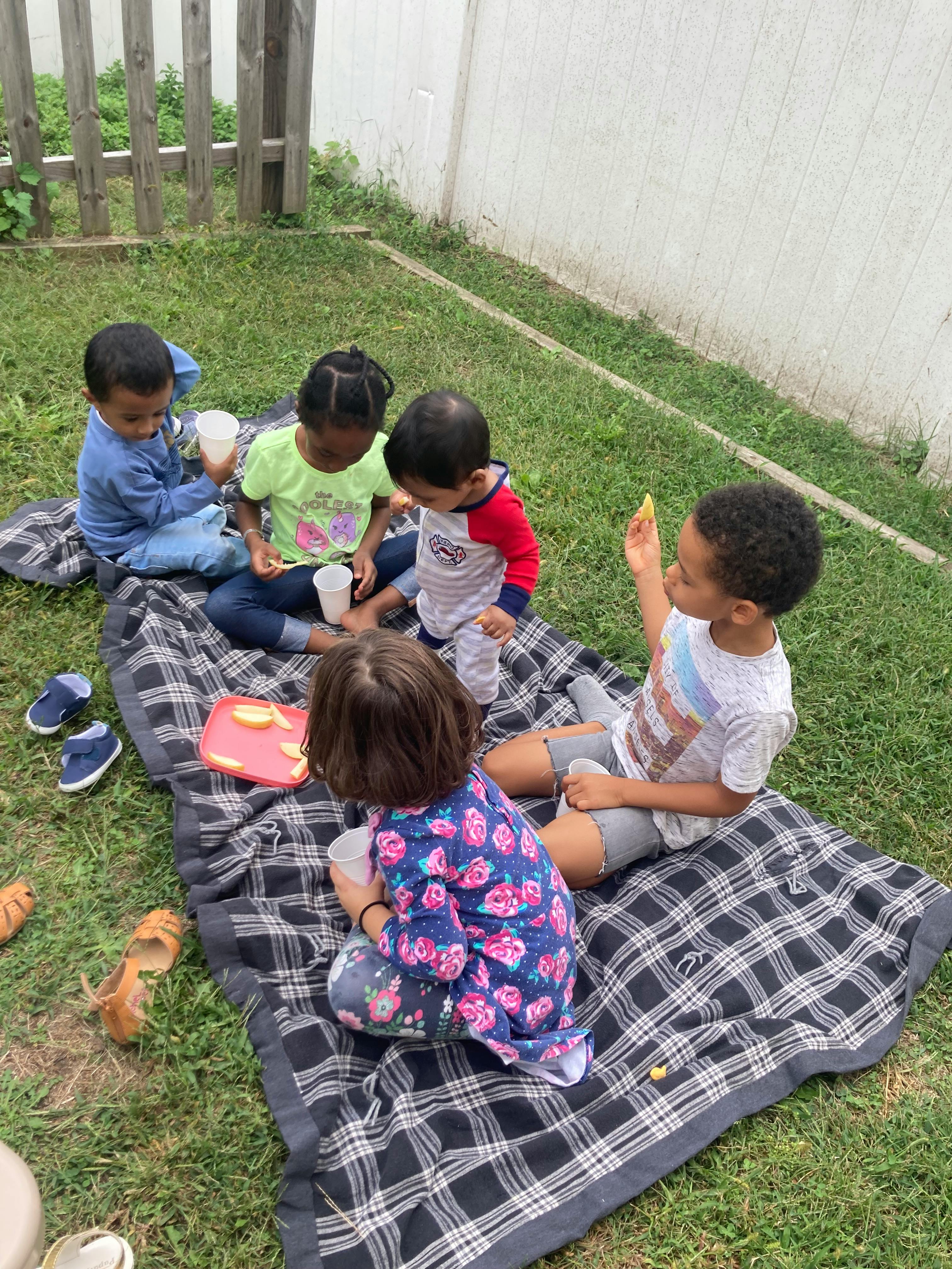 Five children sitting on a plaid blanket on the grass with cups and food
