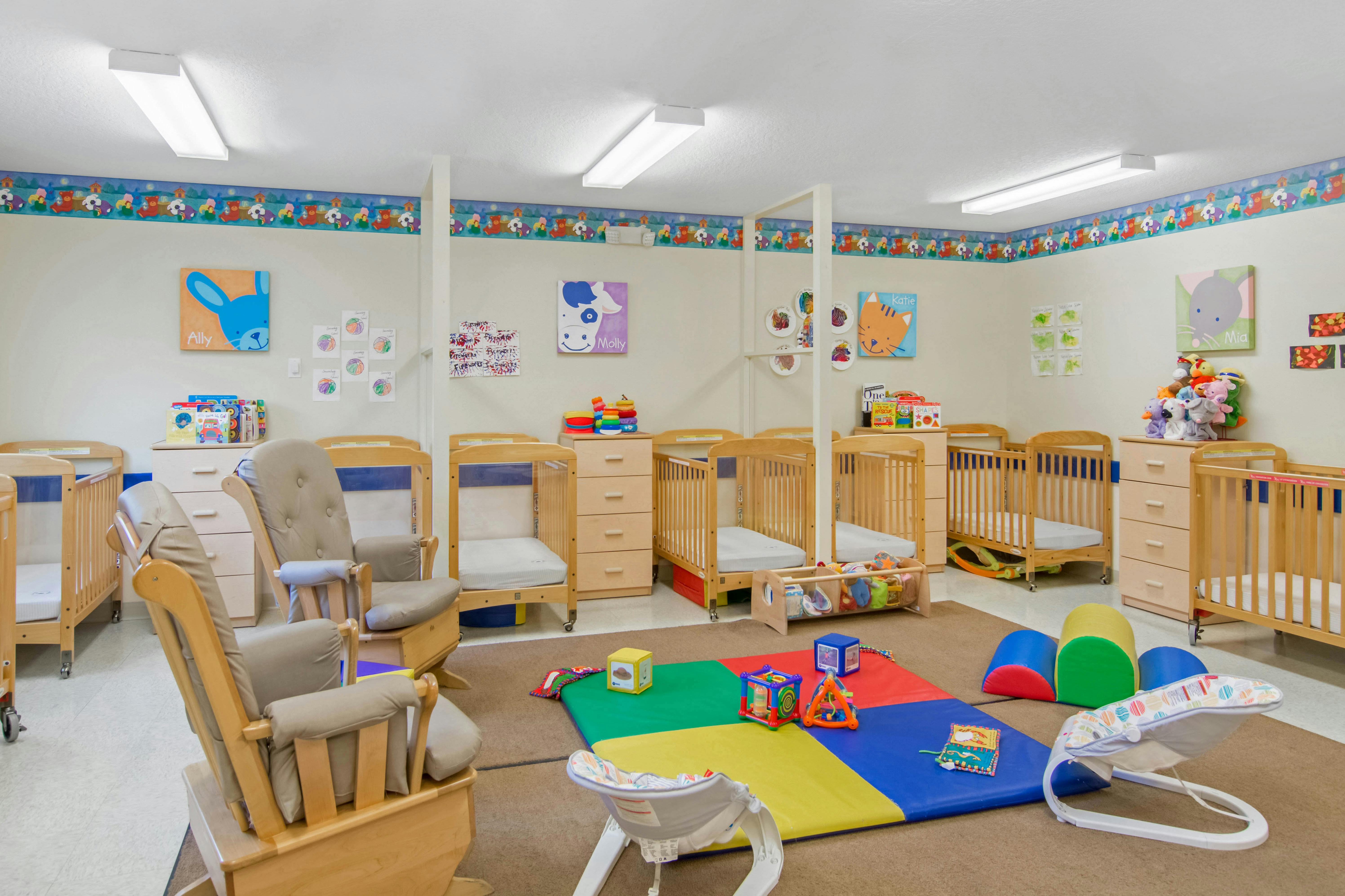 A room with several cribs, dressers, two rocking chairs, and toys on a play mat