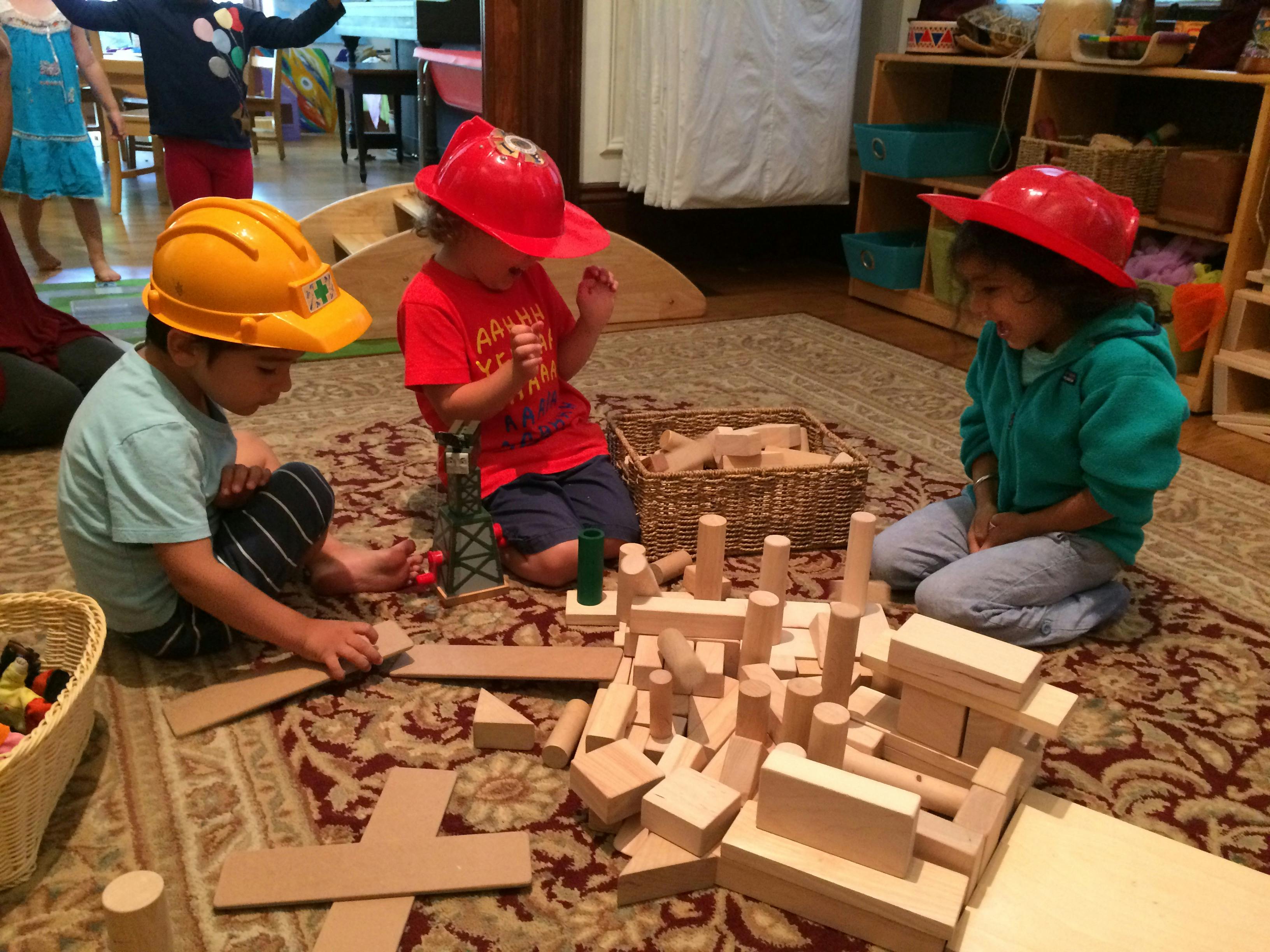 Three children wearing play hats, sitting on a rug and playing with wooden blocks