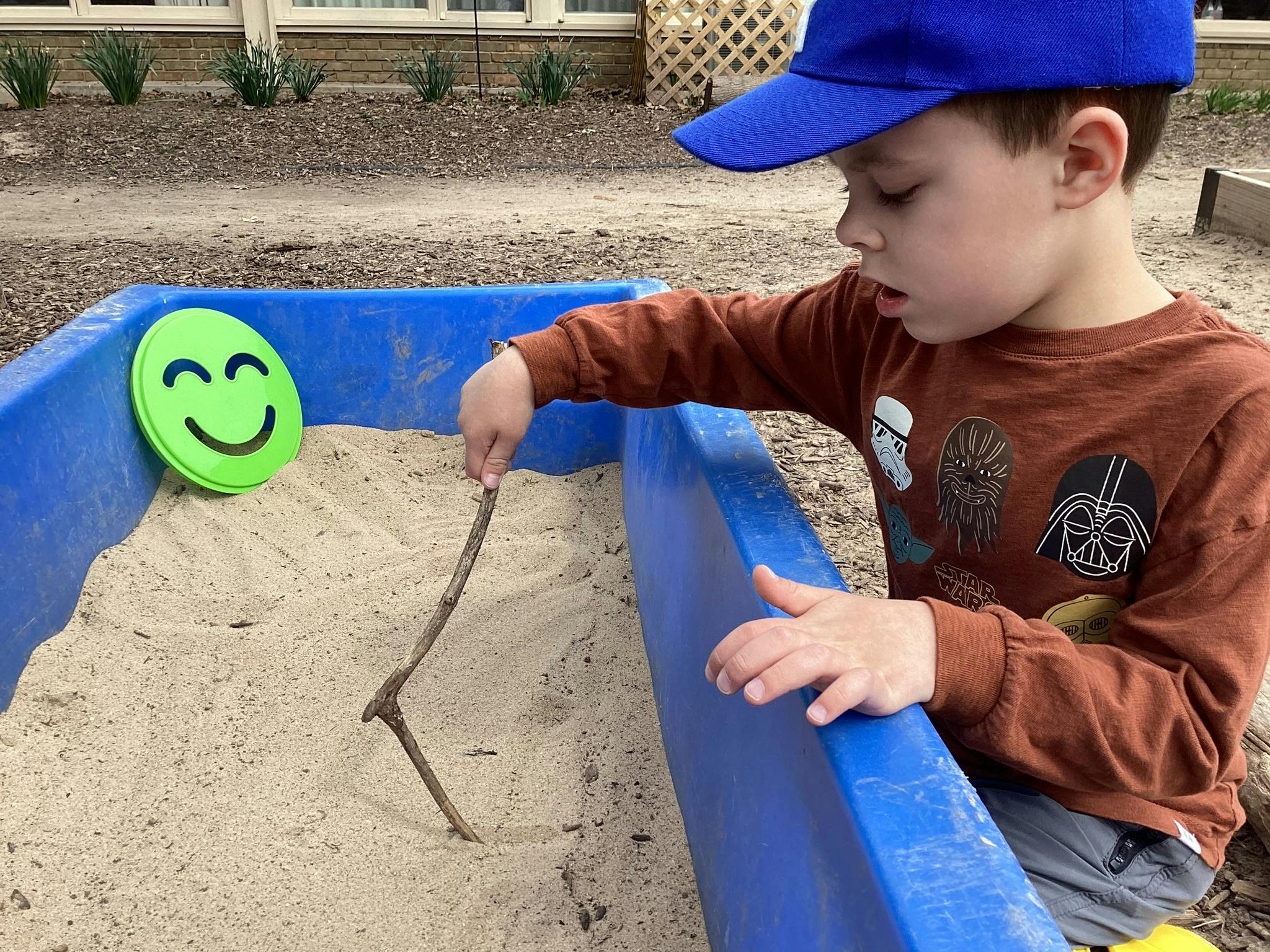 Child in a blue hat playing in a sandbox with a stick