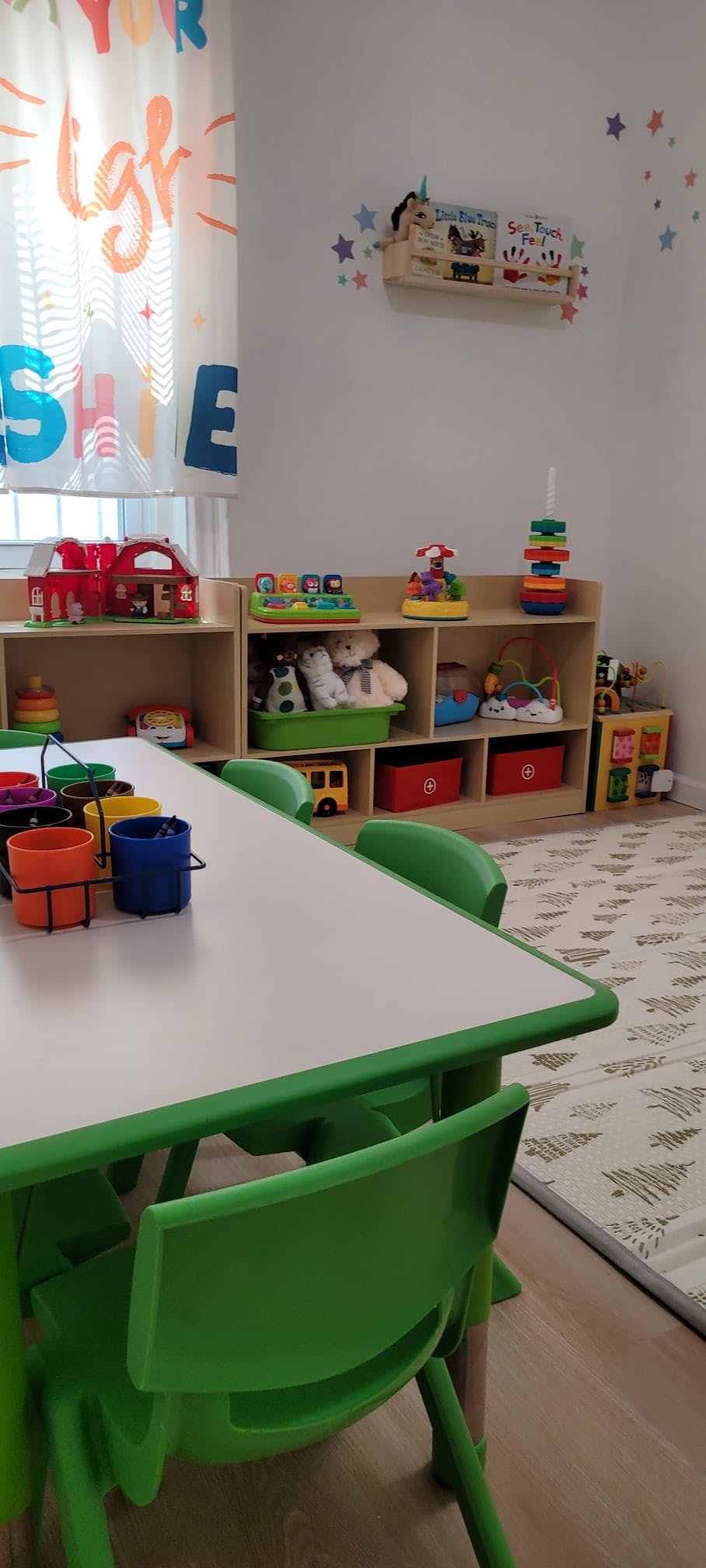 Classroom with green tables and chairs, shelves with toys, and a window with a curtain