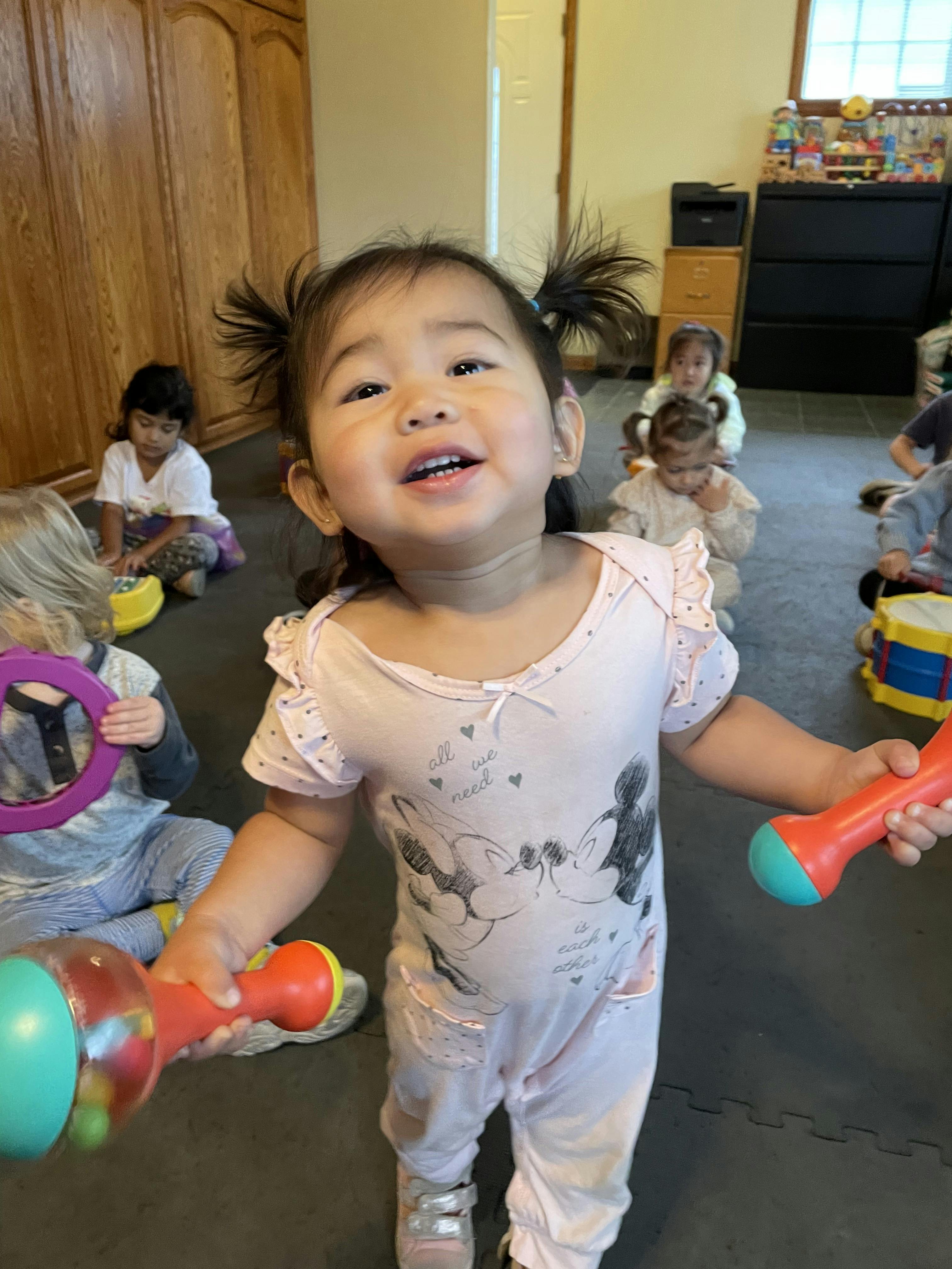 Child holding two colorful maracas in a classroom with other children