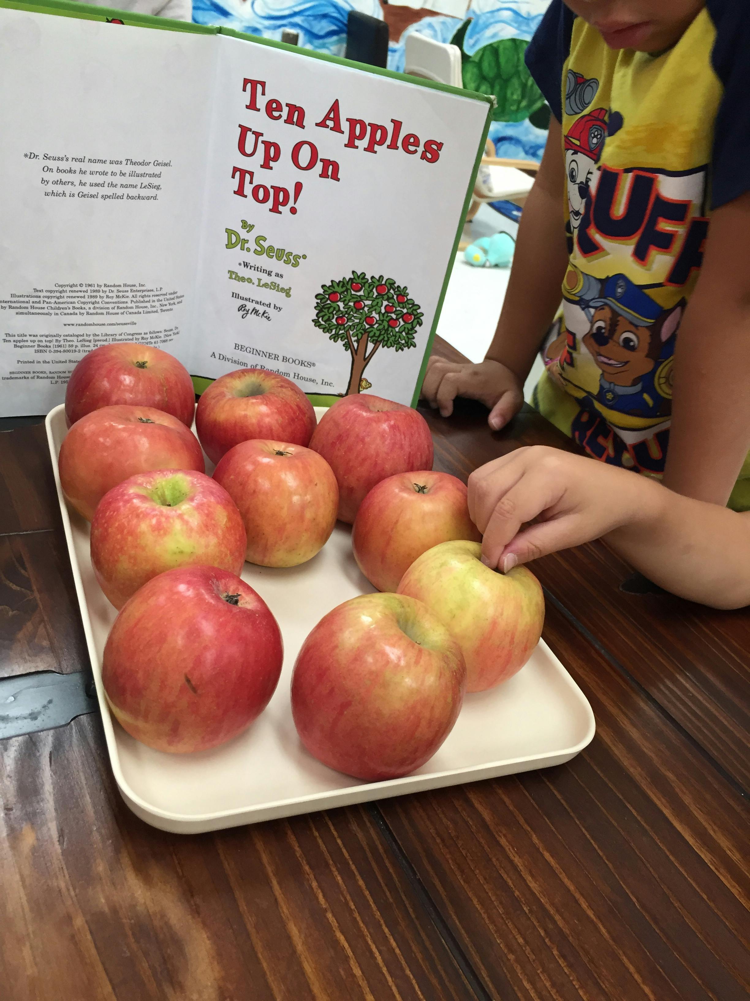 Child reaching for an apple on a tray next to a book titled "Ten Apples Up On Top!"