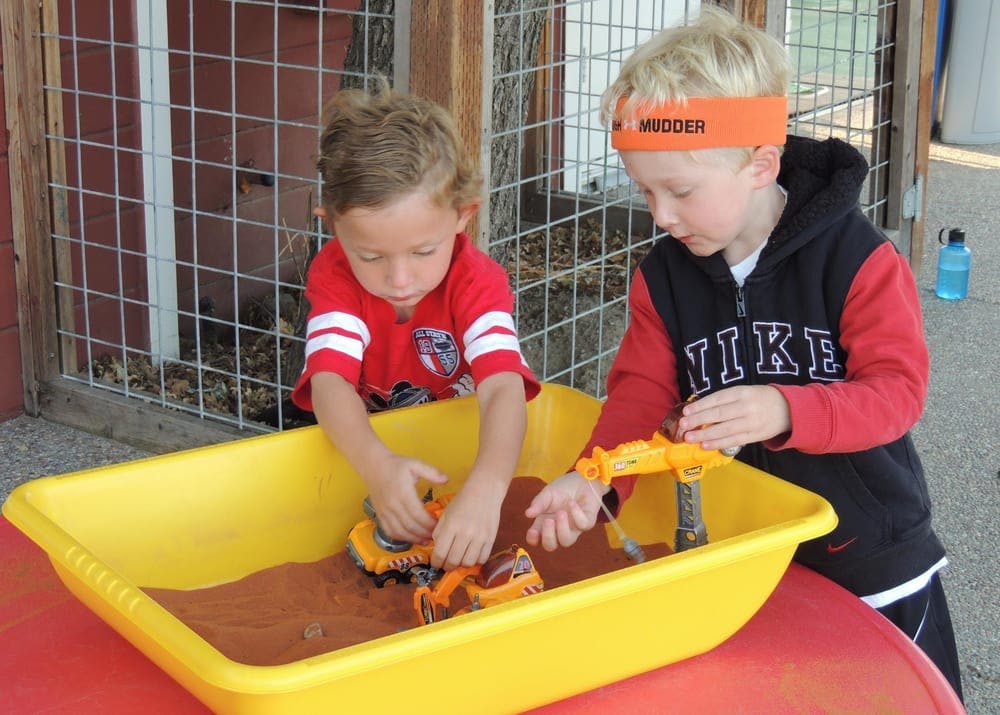 Two children playing with toy construction vehicles in a sand table