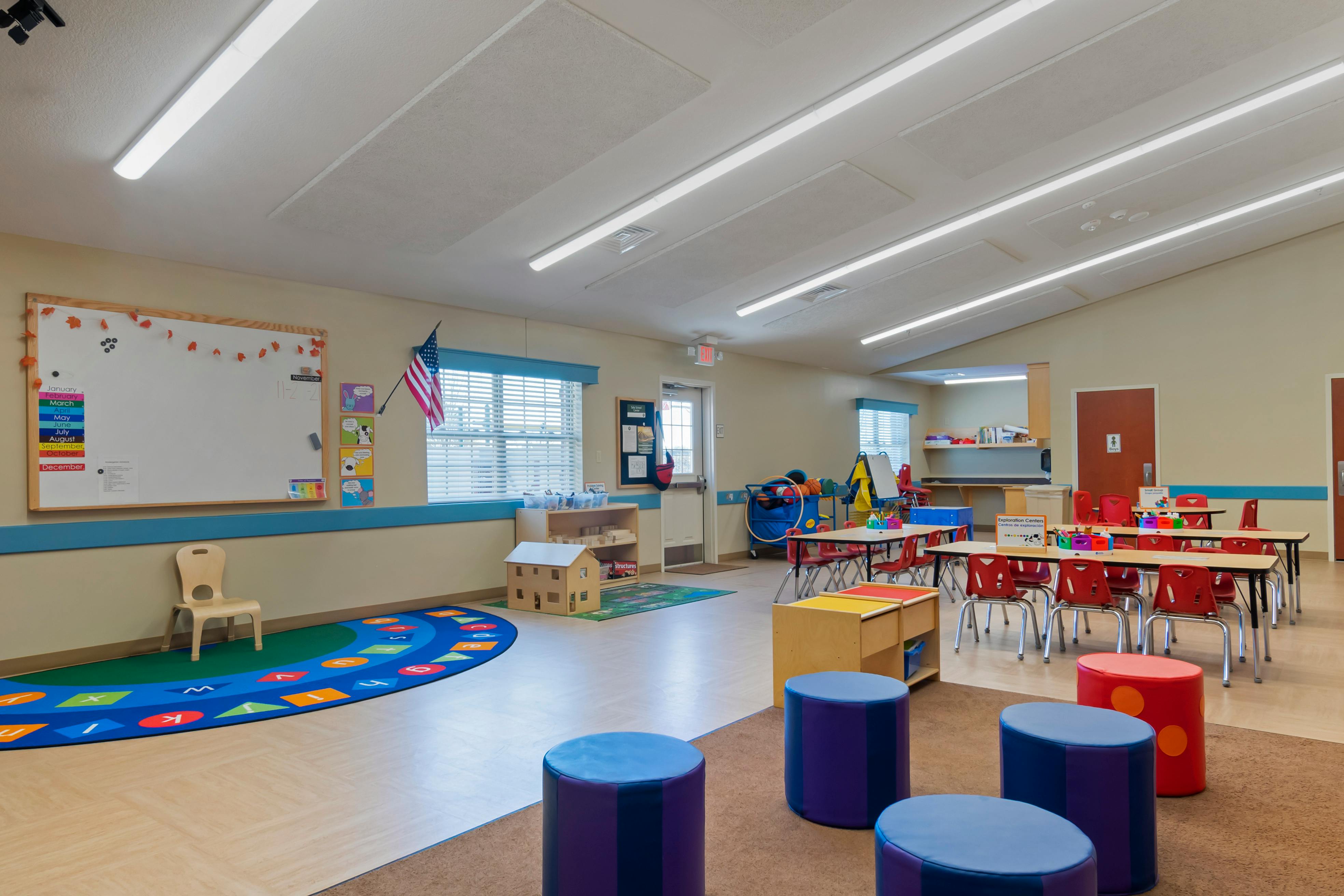 An empty classroom with small tables and chairs, a whiteboard, and play areas