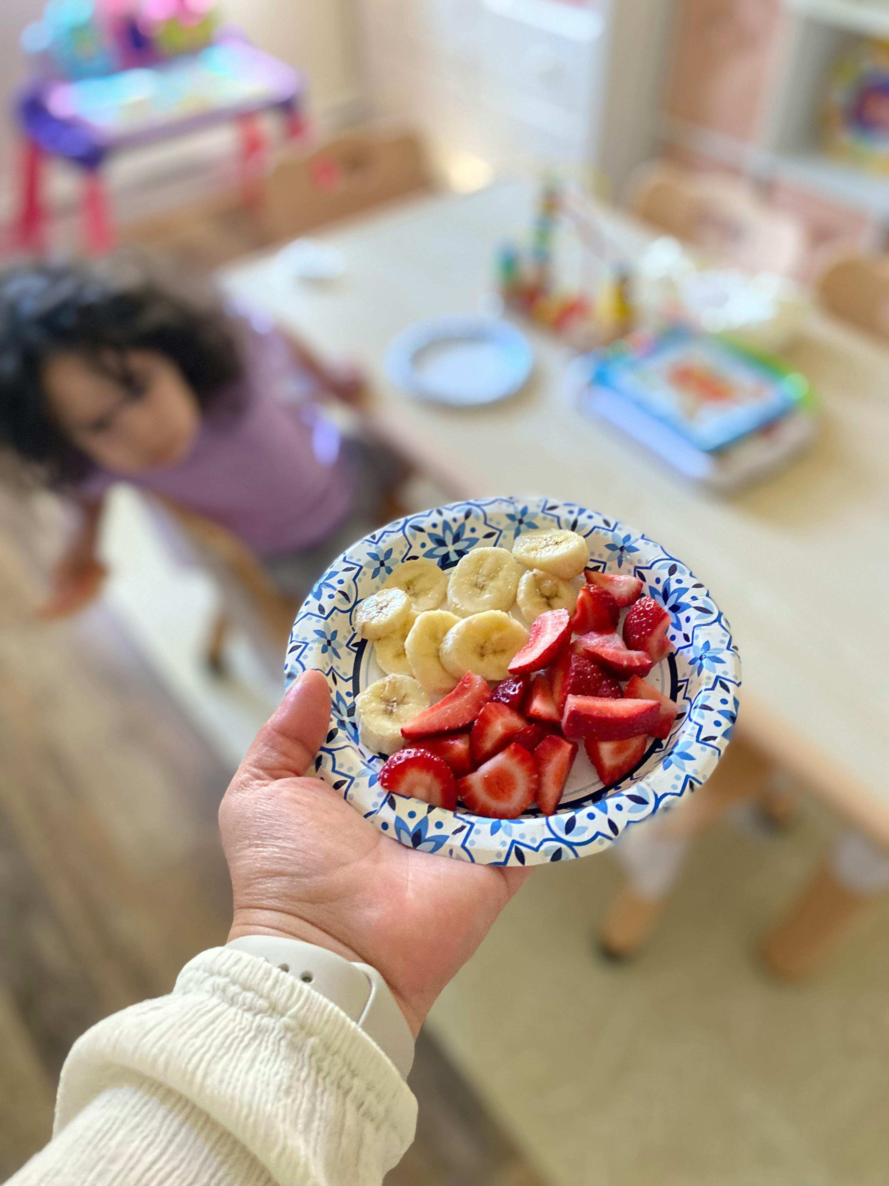 Hand holding a paper plate of sliced bananas and strawberries