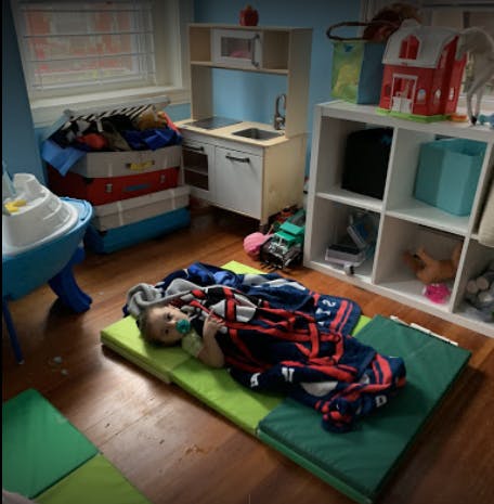 Child lying on a green mat with a blanket in a room with toys