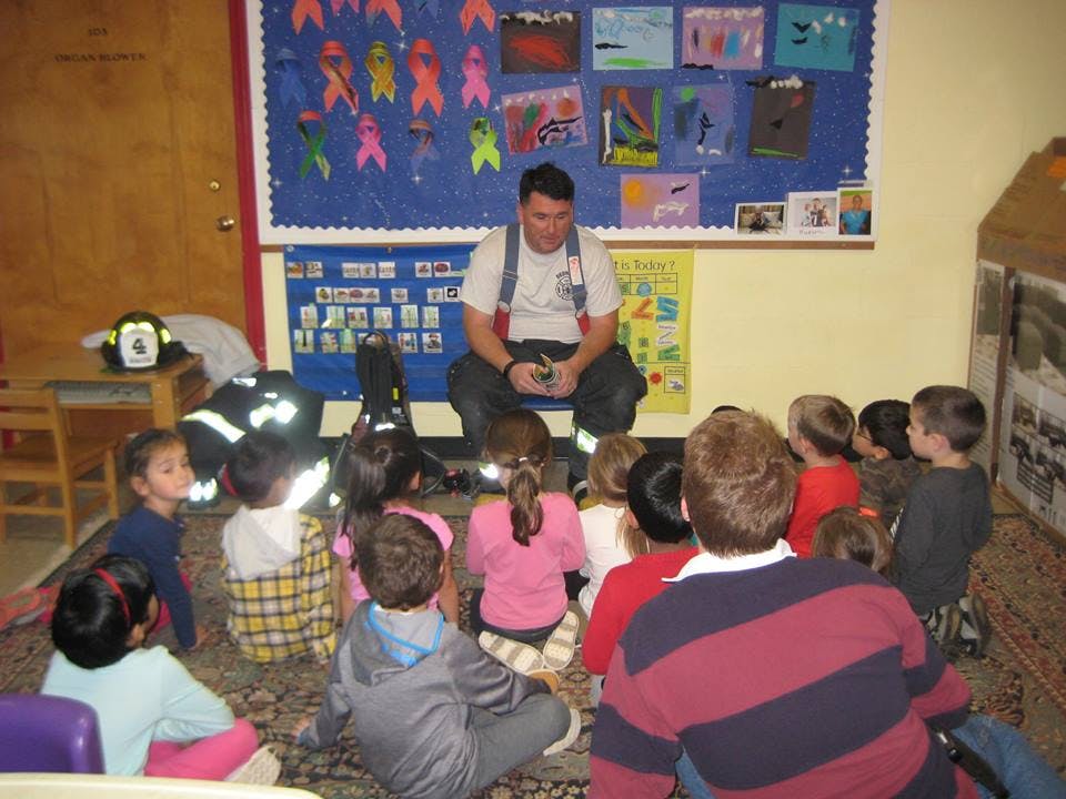 Person in firefighter gear sitting on floor with children in a classroom