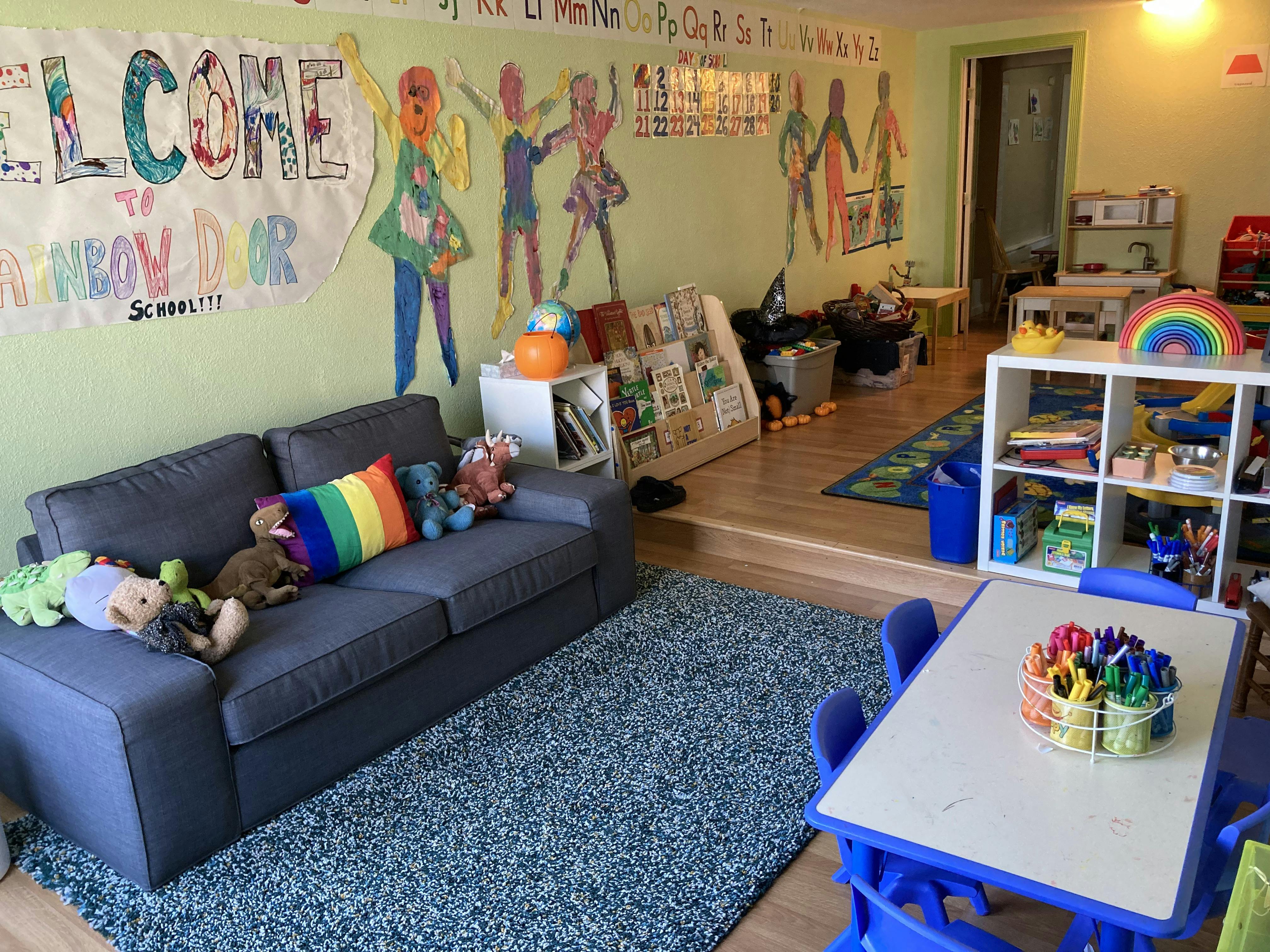 Classroom with sofa, play kitchen, small table, and "Welcome to Rainbow Door School" sign