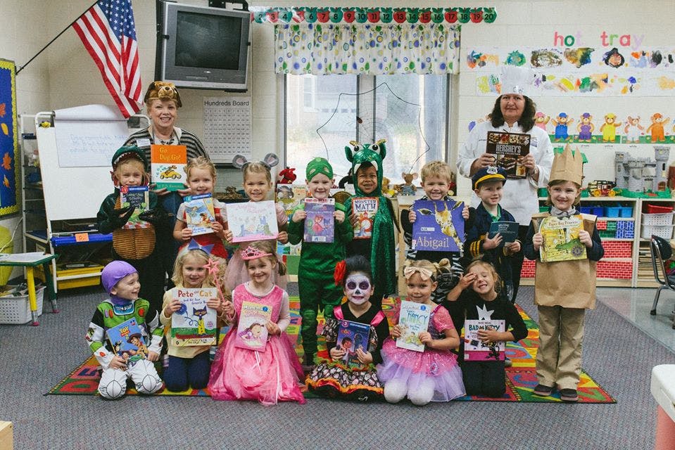 Two adults and multiple children in costumes holding books in a classroom