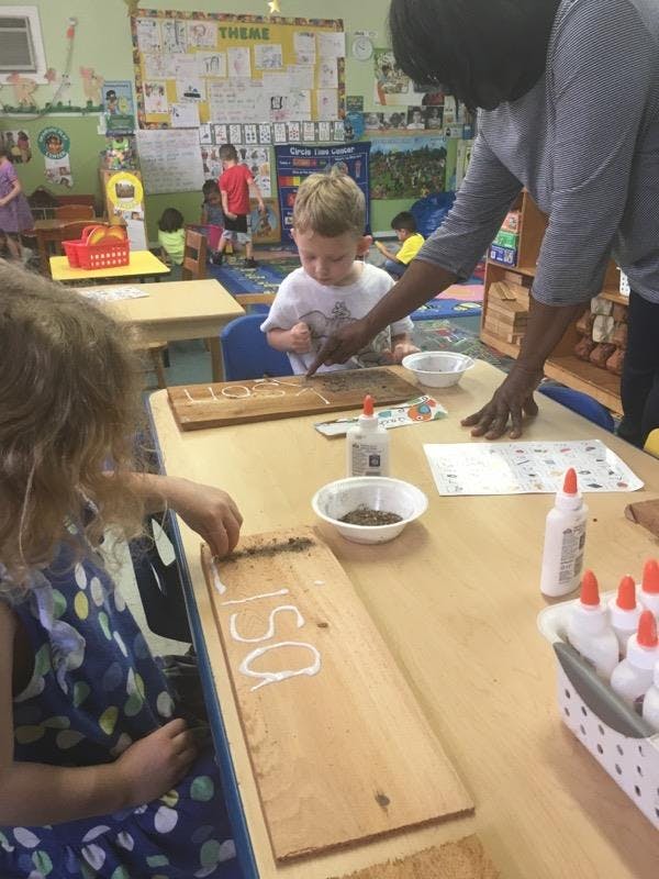 Adult helping a child with a craft project at a table in a classroom