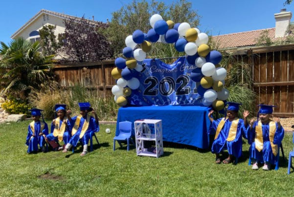 Children in graduation gowns sitting on grass in front of a balloon arch
