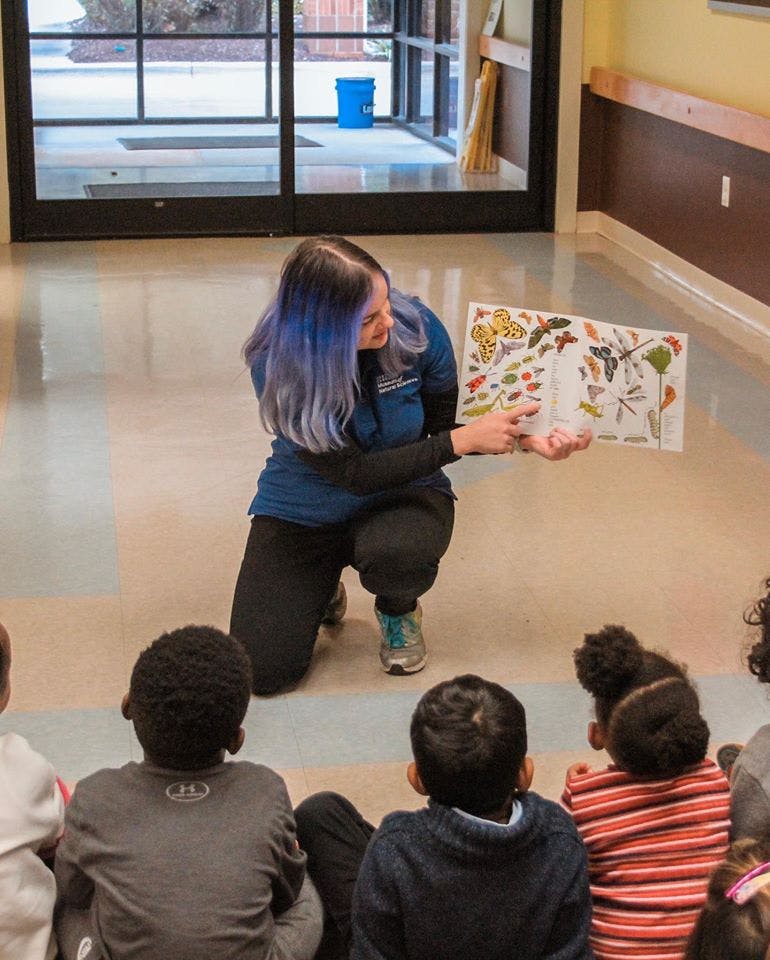 Person kneeling and showing a book to children sitting on the floor