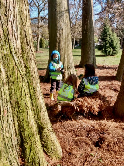 Three children in safety vests playing outdoors near tree trunks