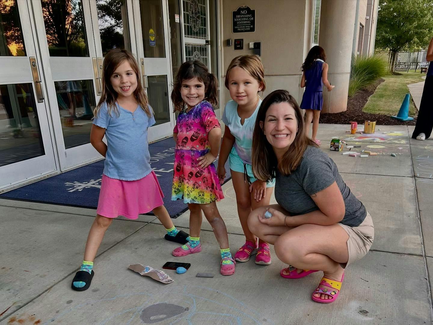 Three children and one adult squatting near chalk drawings on a sidewalk