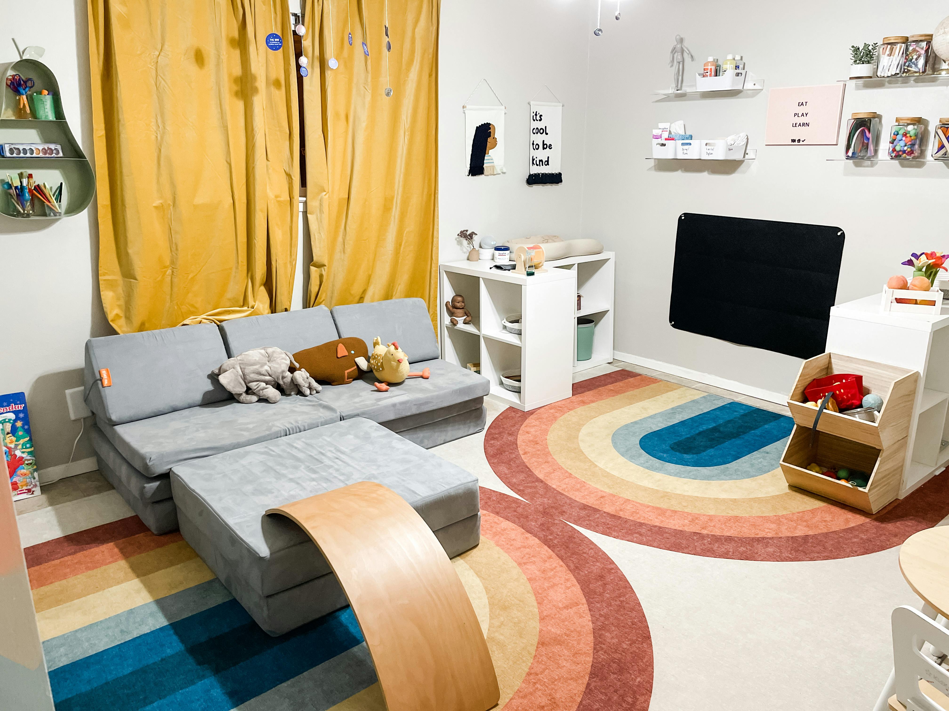 Playroom with a gray modular sofa, rainbow rug, and white shelves