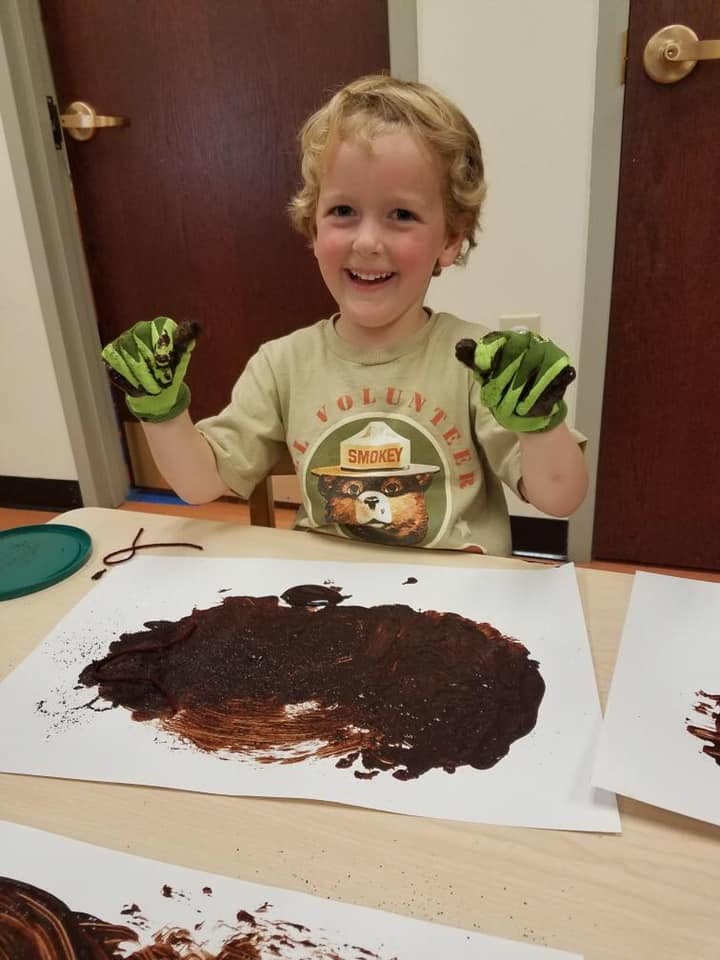 Child wearing gloves and a Smokey Bear shirt, finger painting with brown paint