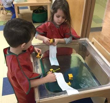 Two children playing with toy trucks and paper in a water table