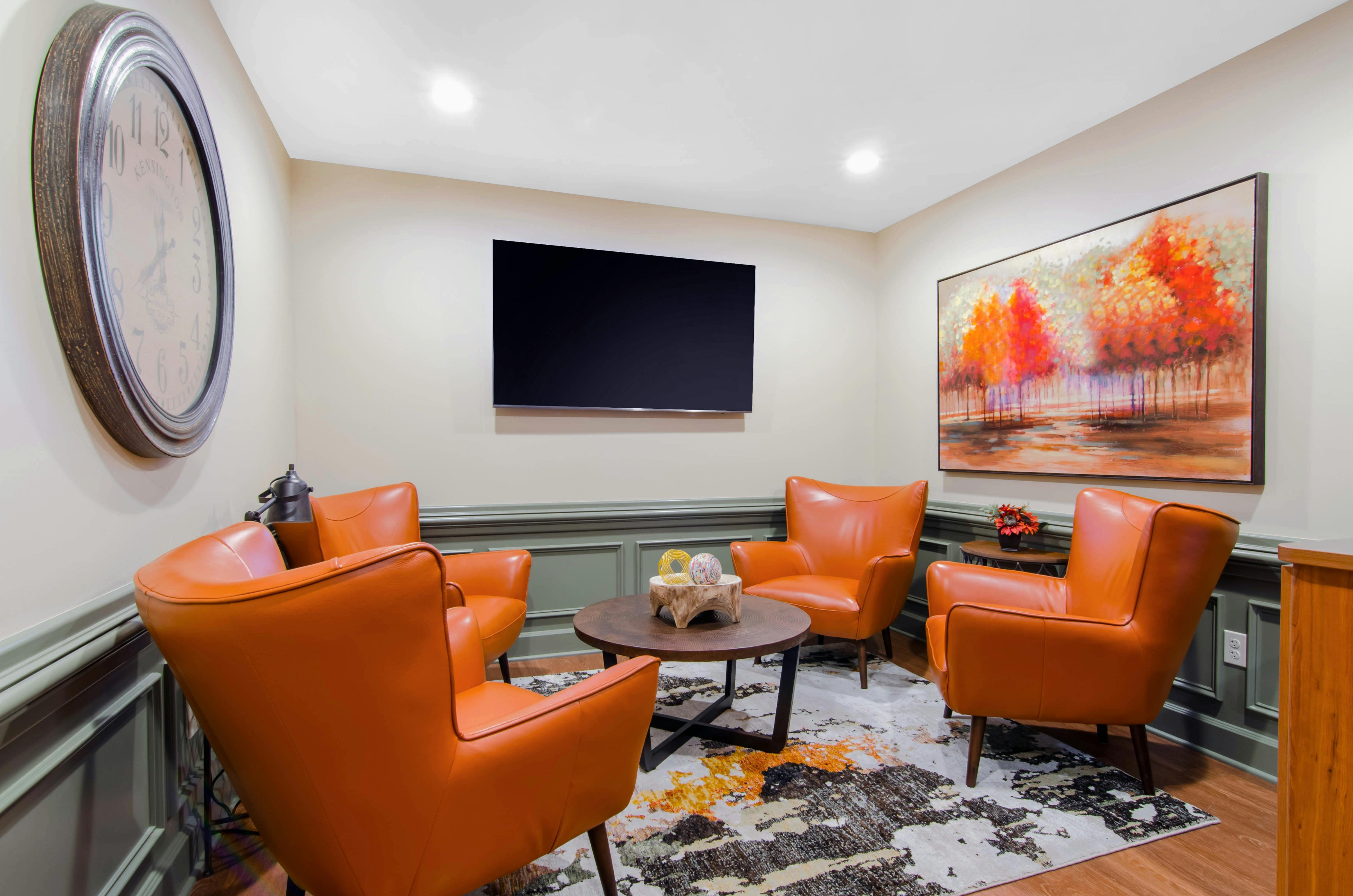 Four orange chairs around a coffee table with a large clock and TV on the walls