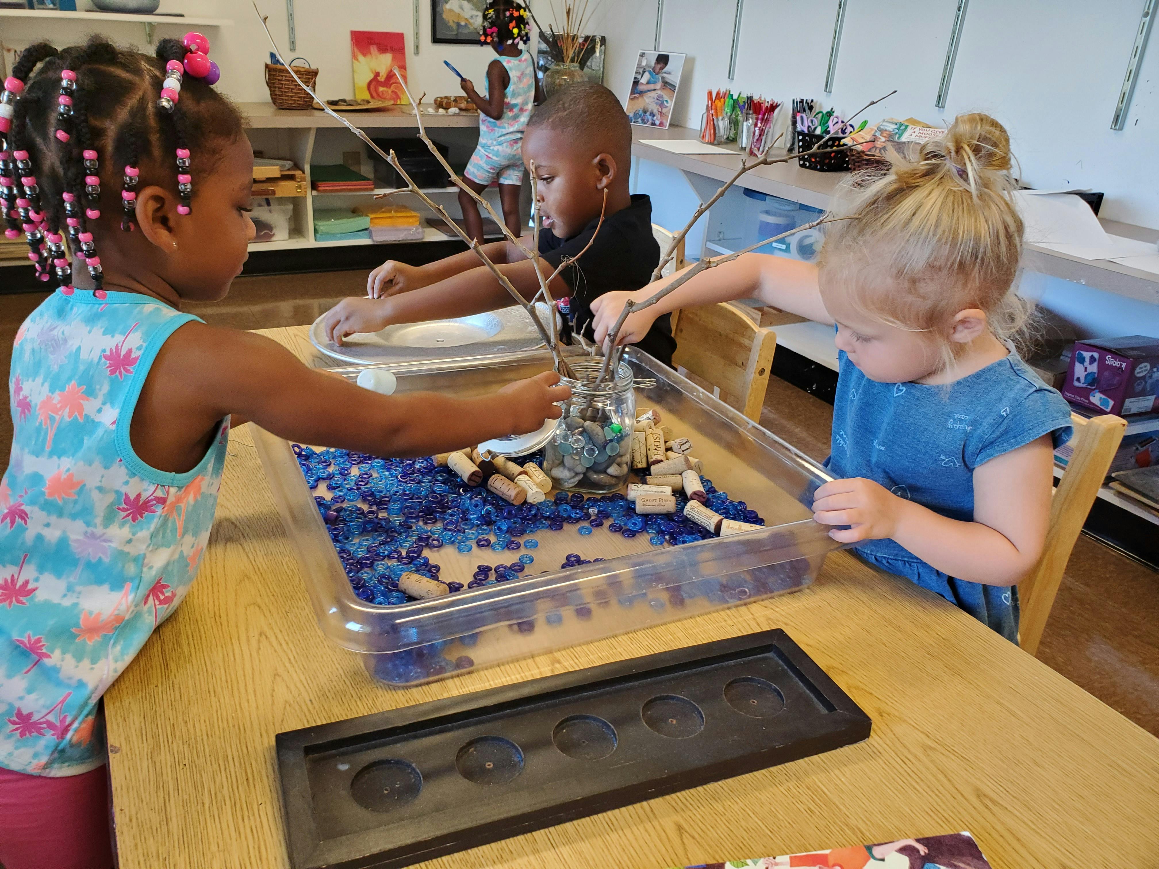 Three children playing with blue beads and sticks in a clear bin