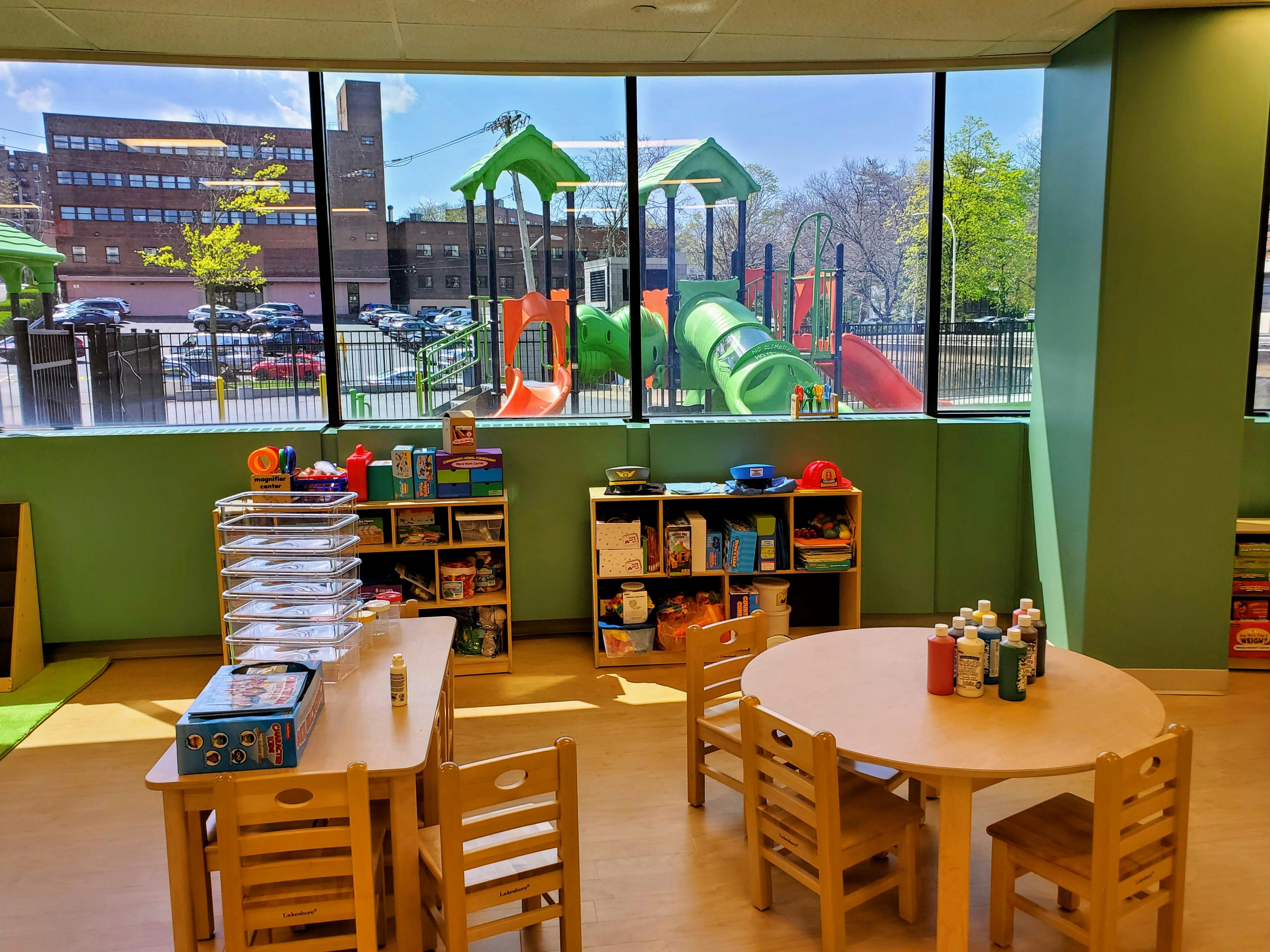 Indoor classroom with tables and chairs, outdoor playground visible through window