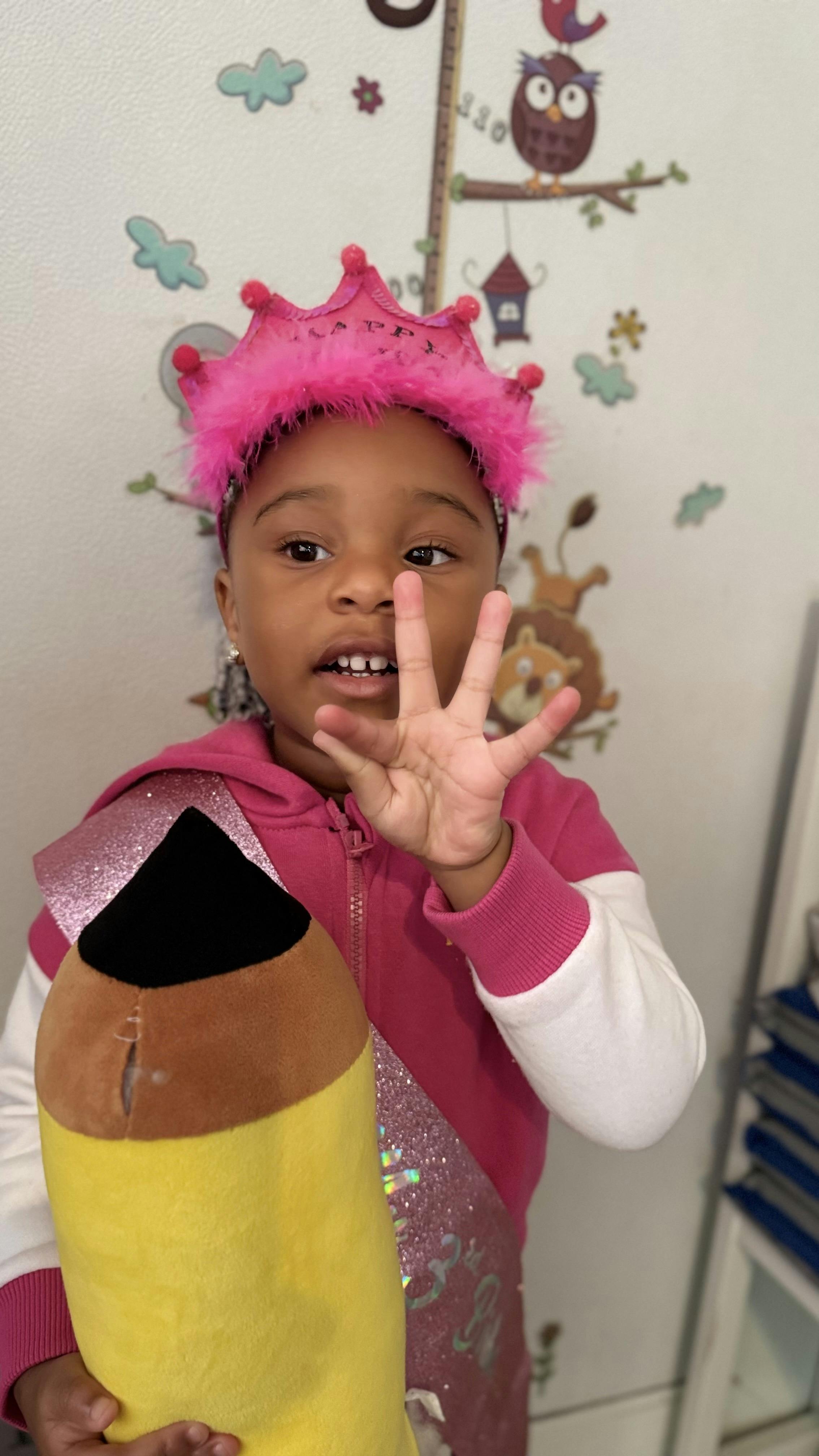 Child wearing a pink crown holding up four fingers and a plush pencil