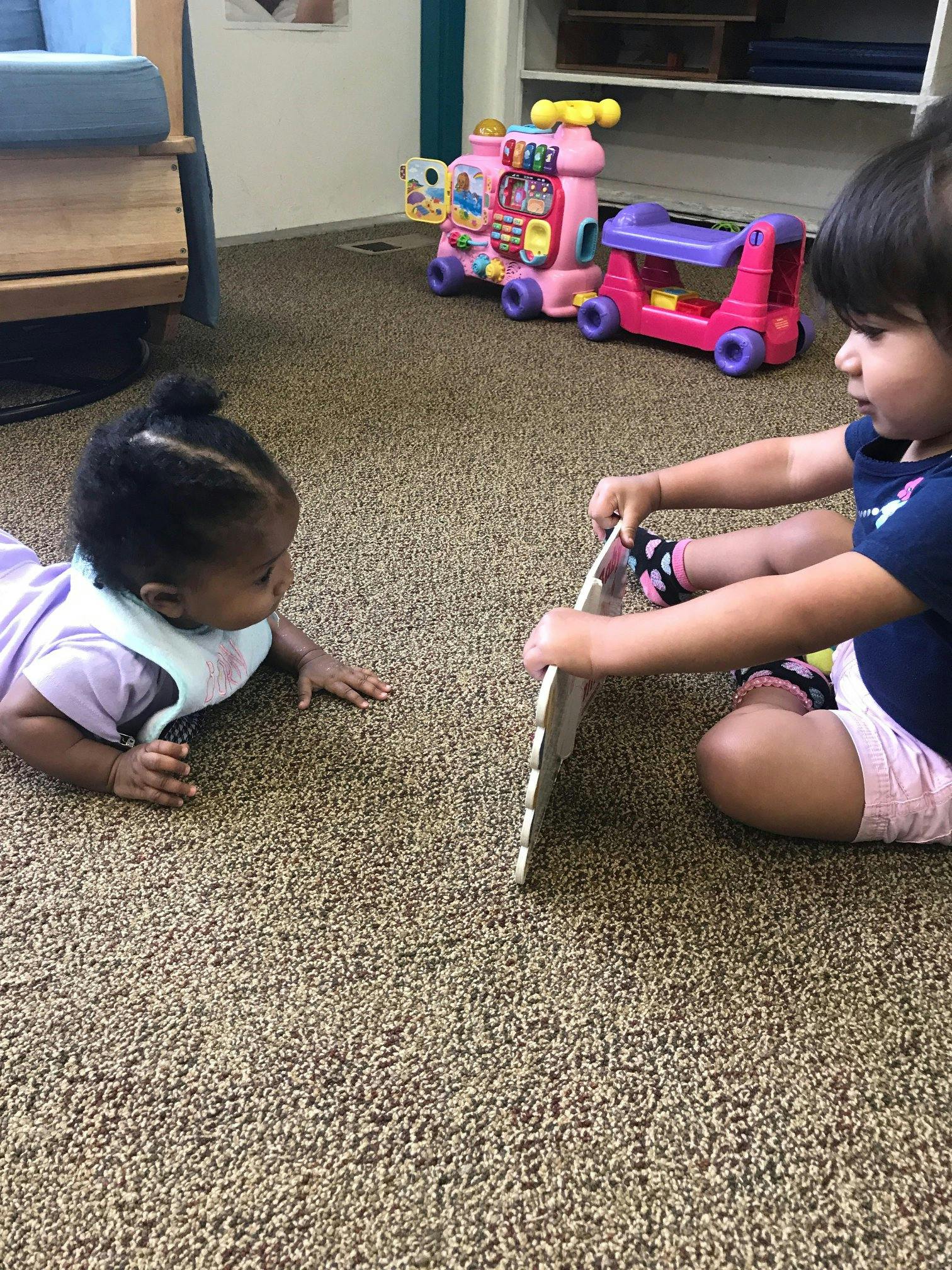 Child on floor holding a toy, another child on stomach looking at toy
