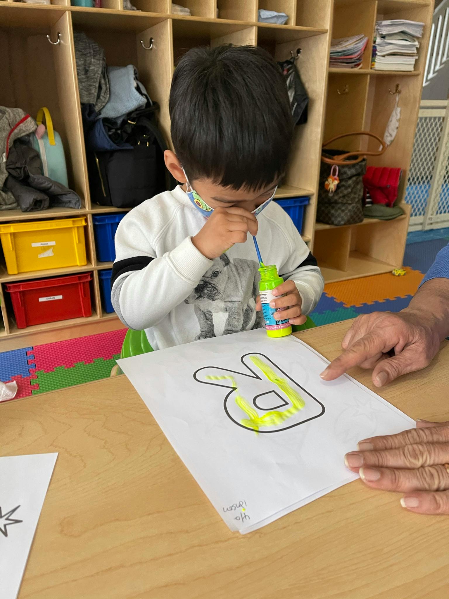 Child coloring a letter on paper with a person's hand pointing