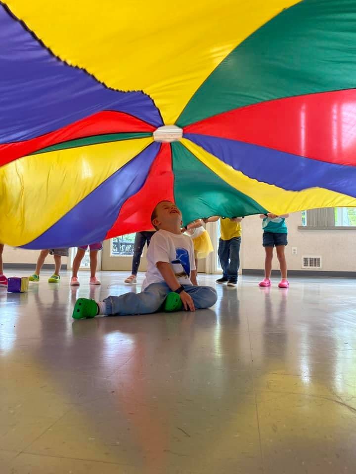 Child sitting on floor under a colorful parachute with other people standing