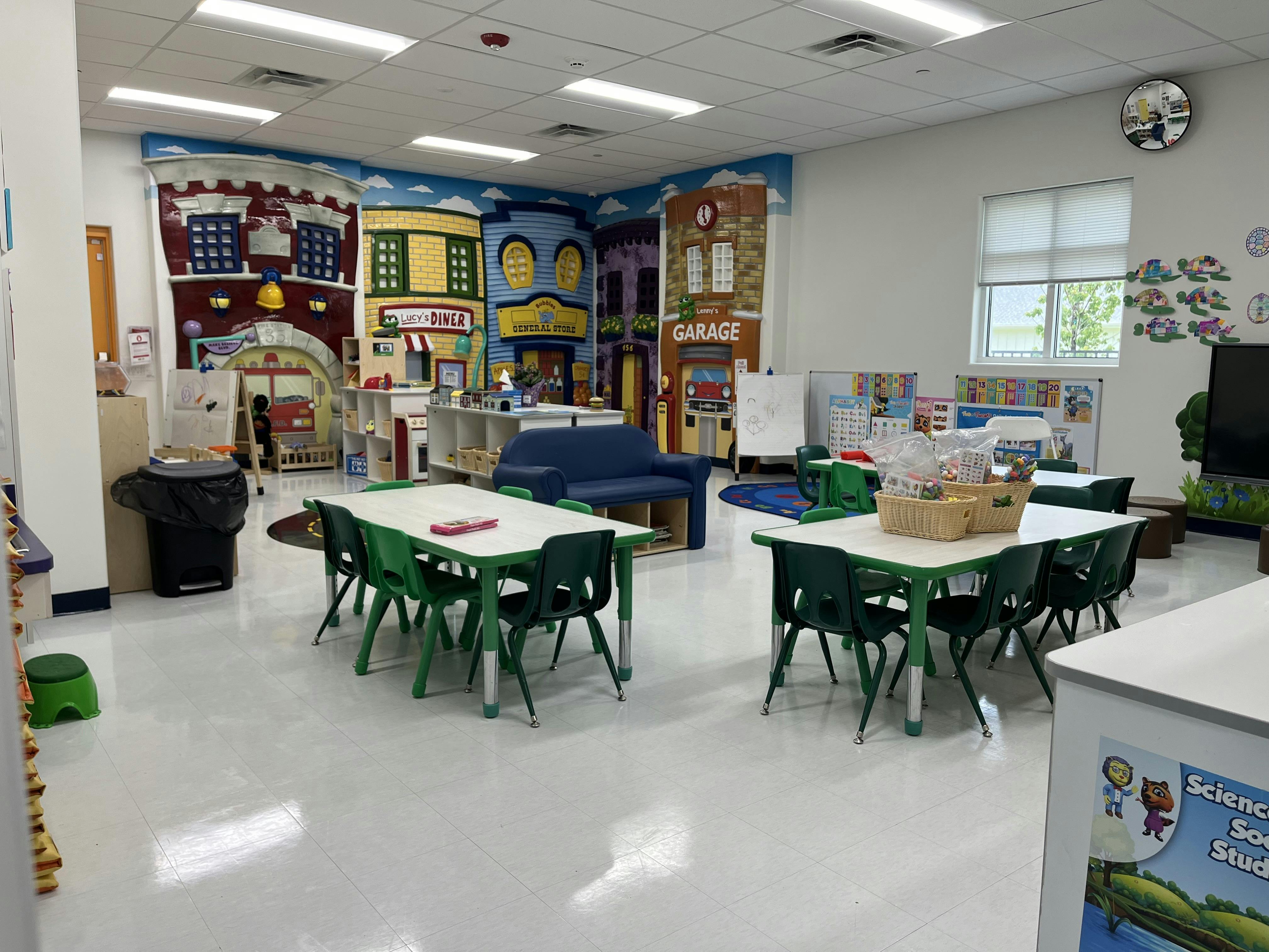 Empty classroom with small tables and chairs, and a play area with a city mural