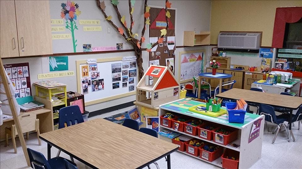 Classroom with tables, chairs, a dollhouse, and learning displays on the walls