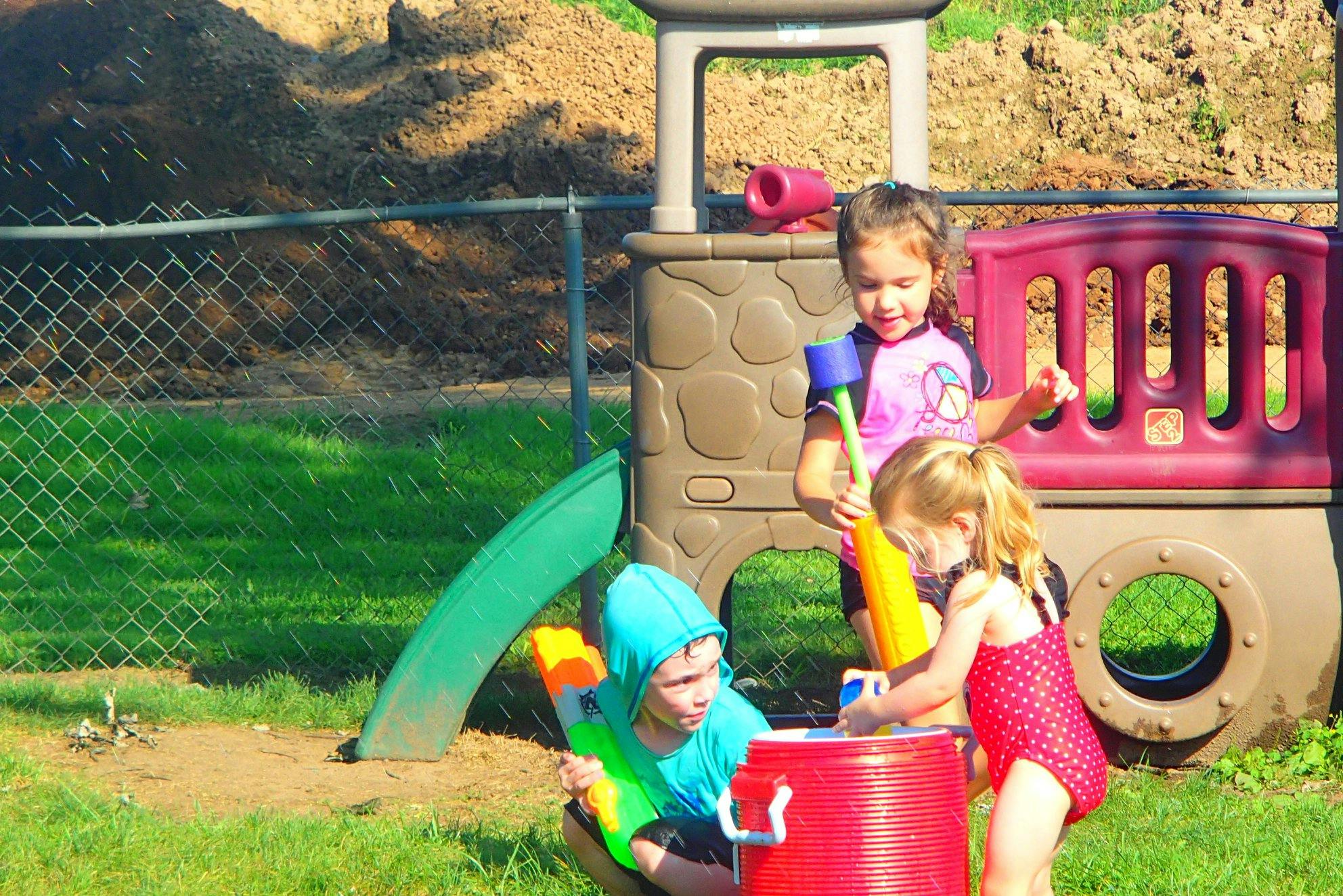 Three children playing with water toys and a red bucket outdoors