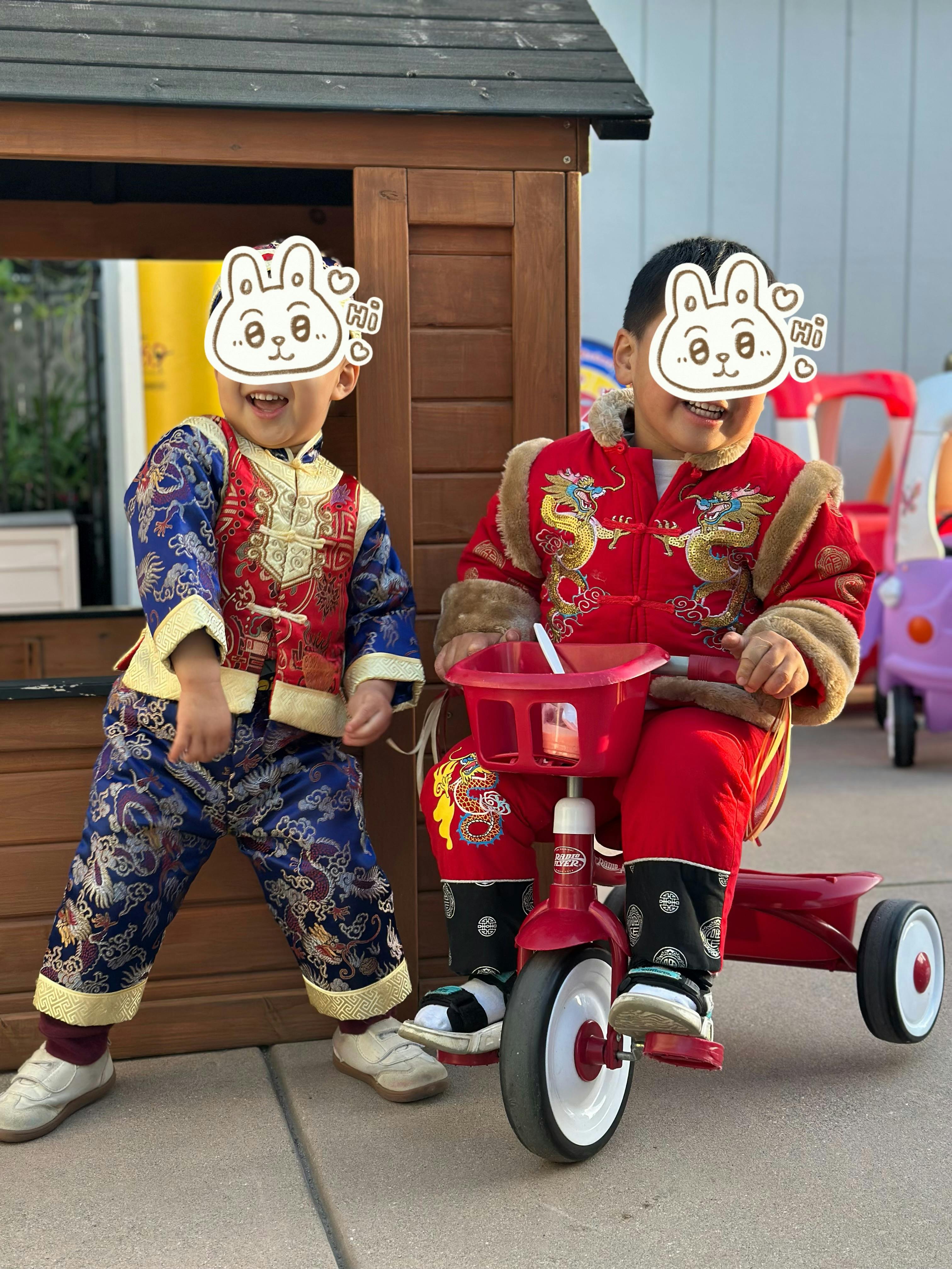 Two children wearing traditional clothing, one standing and one on a tricycle