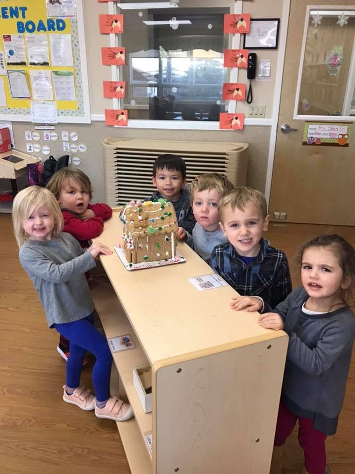 Six children standing around a table with a gingerbread house