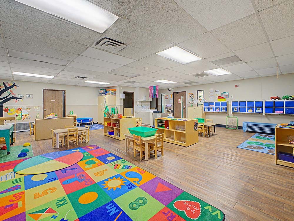 Colorful classroom with small tables and chairs, shelves, and a large alphabet rug
