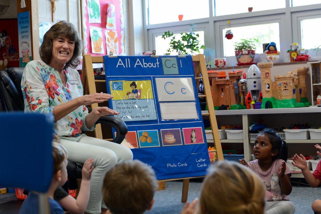 Person sitting in front of children in a classroom, gesturing toward a board