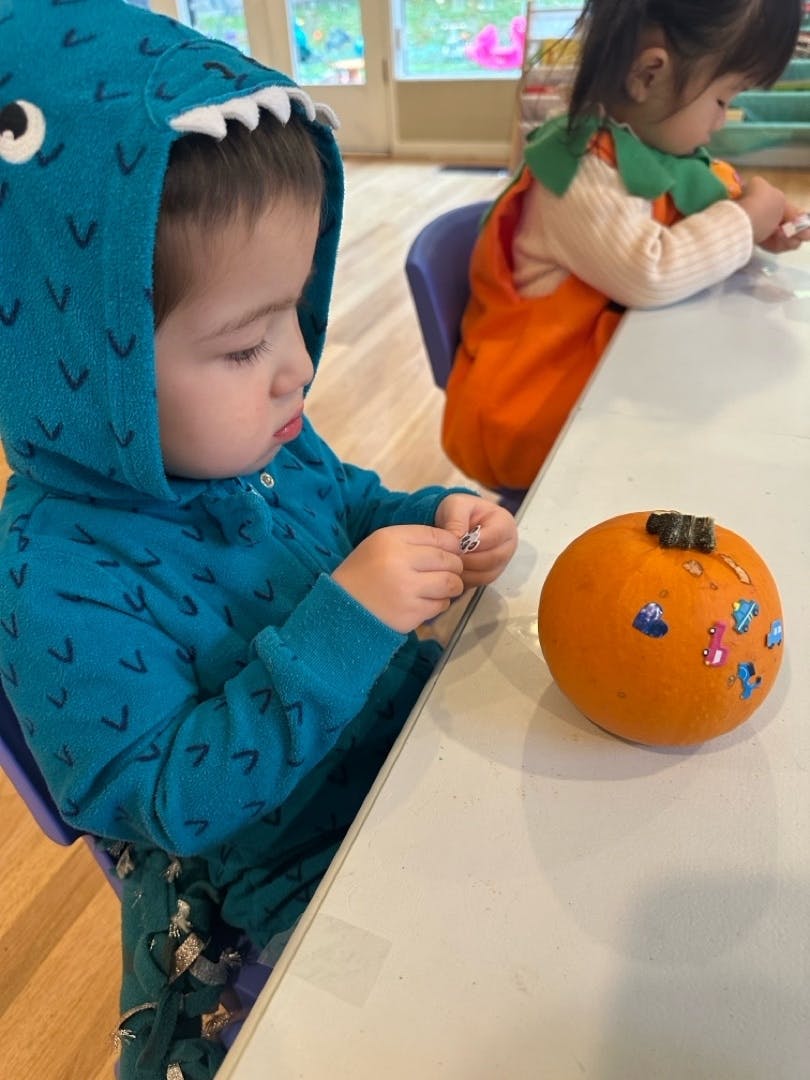 Child in dinosaur costume decorating a pumpkin with stickers, another child in pumpkin costume
