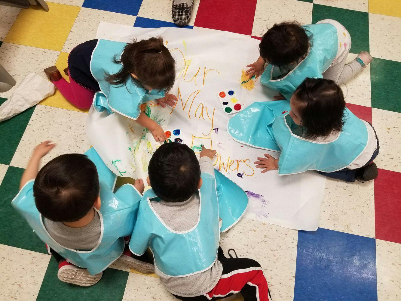 Five children in smocks painting on a large sheet of paper on the floor