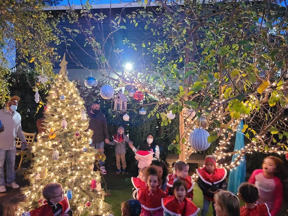 People and children gathered around a lit Christmas tree and string lights