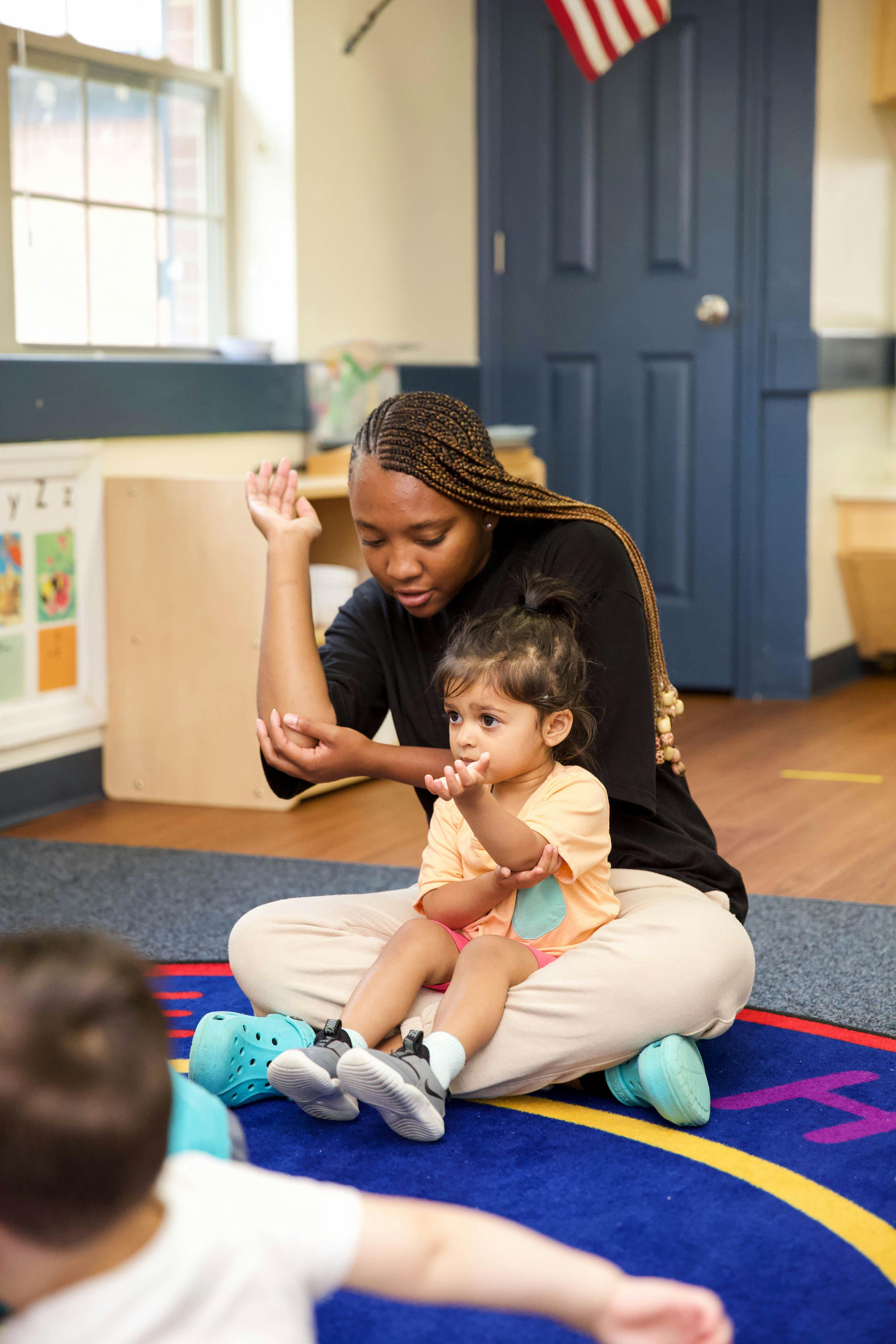 Person on floor with a child in a classroom, both raising their hands
