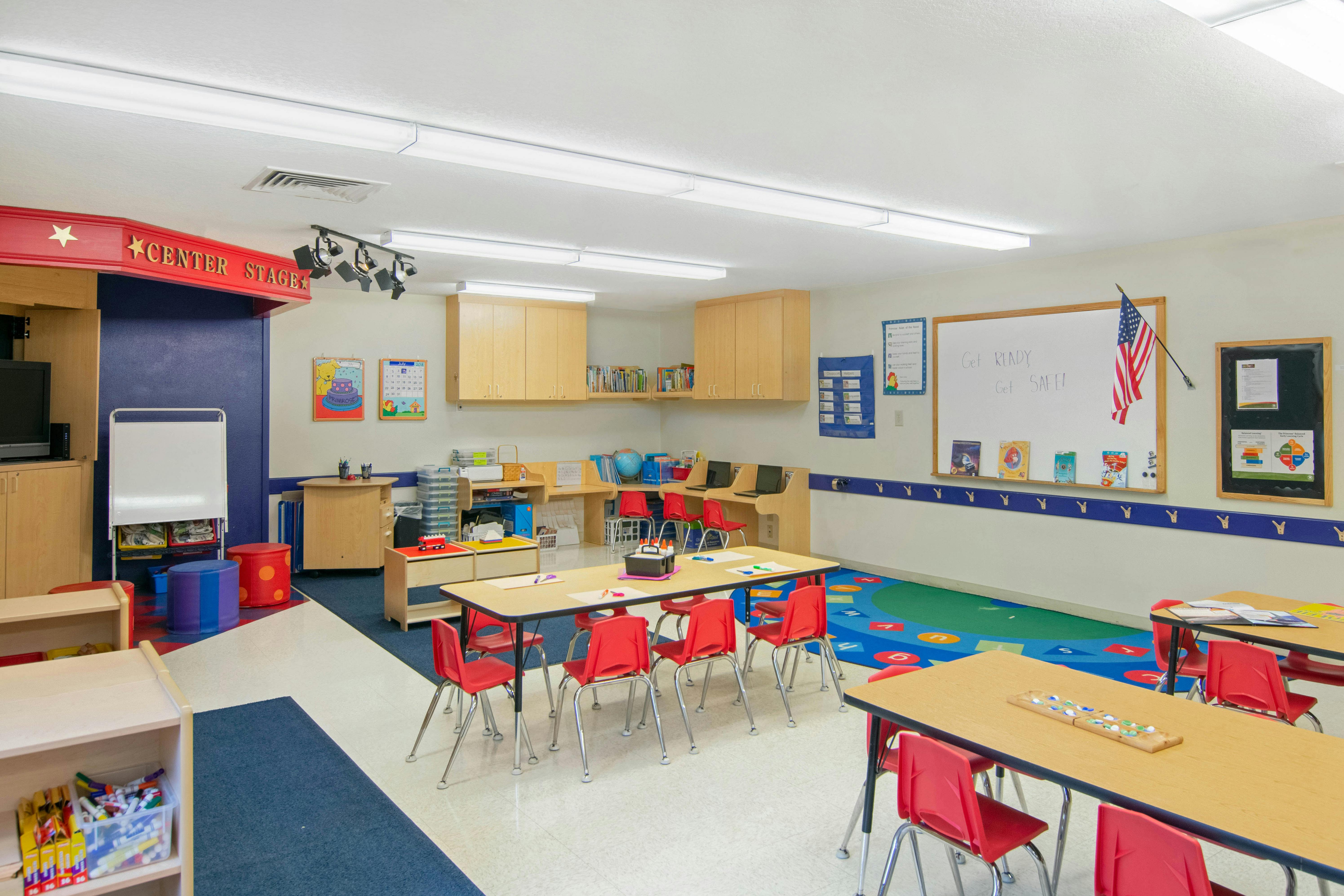 Empty classroom with small red chairs and tables, a whiteboard, and a stage